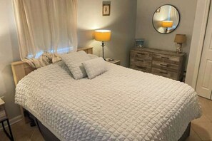 Une chambre à coucher comprenant un lit, une table de chevet, une commode, une lampe, un miroir et une fenêtre avec des rideaux.