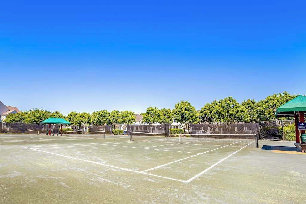 An outdoor tennis court with a net, green umbrellas, and trees in the background.