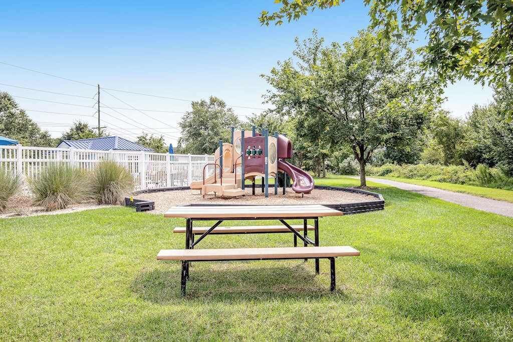 A playground with a slide and climbing structure, surrounded by a white fence and trees.
