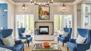 A living room with a fireplace, blue armchairs, a coffee table with books and a bowl, and a painting above the fireplace. - Millville by the Sea home with clubhouse, pools & fitness center access (Millville)