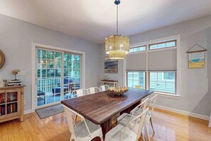A dining area with a wooden table, white chairs, a hanging light fixture, and large windows. - Ocean Mist townhouse with deck, grill, bikes, WIFI & W/D (Ocean View)