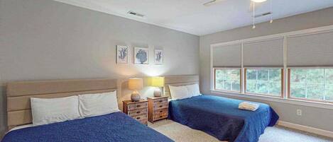 A hotel room with two beds, a ceiling fan, a window with blinds, and a nightstand with a lamp.