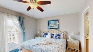 Une chambre à coucher comprenant un lit, un ventilateur de plafond, une table de chevet, un banc, une fenêtre avec des rideaux et un tableau encadré accroché au mur.