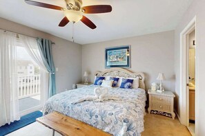 A bedroom with a bed, a ceiling fan, a nightstand, a bench, a window with curtains, and a framed picture on the wall.