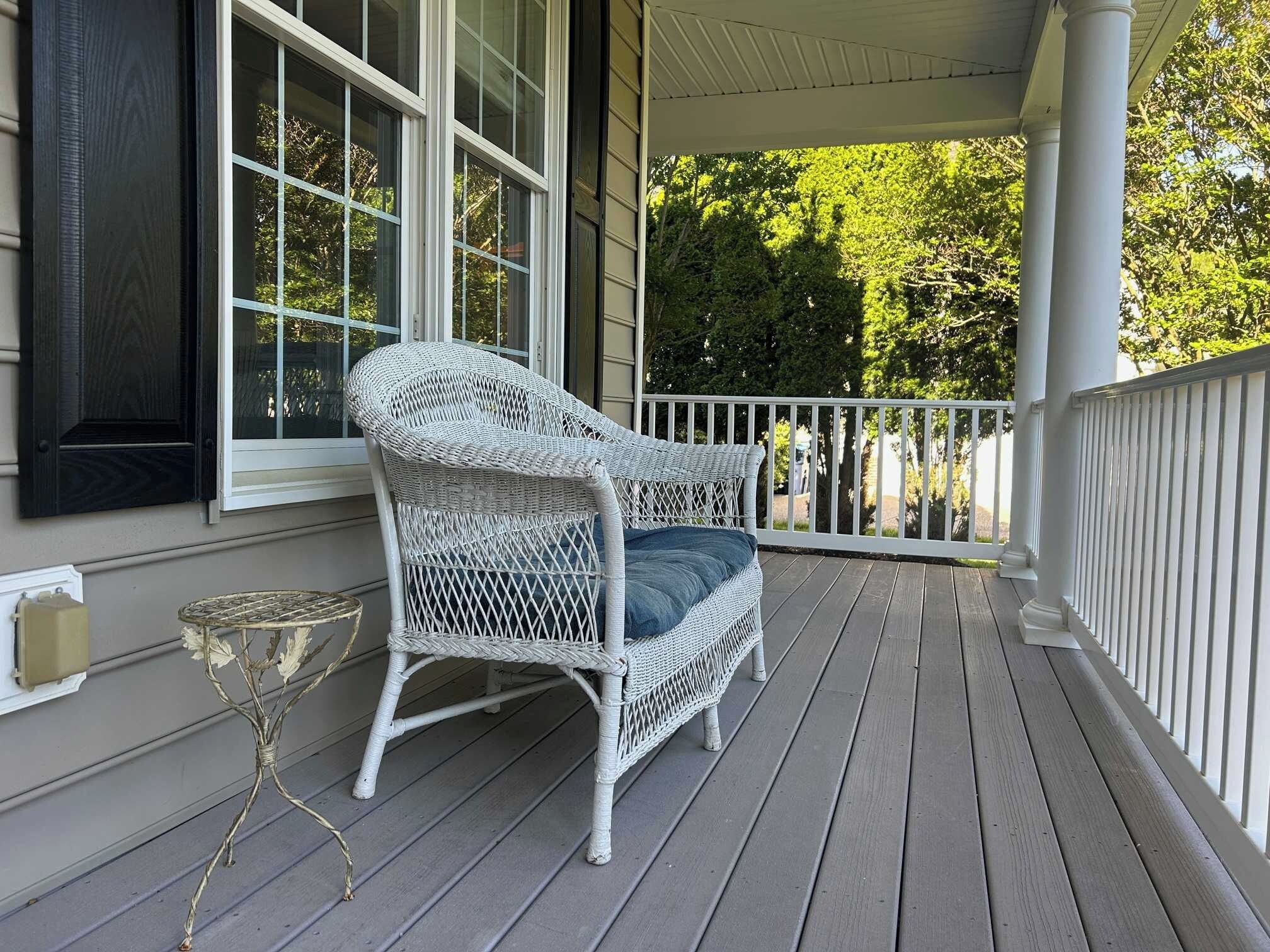 A white wicker chair with a cushion on a wooden porch with a white railing.