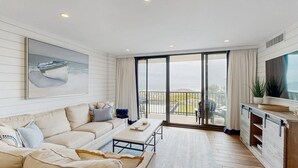 A modern living room with a sofa, coffee table, and a large TV. There is a balcony with a view of the sea. - Sea Colony 2nd floor oceanside condo w/community pools, tennis, fitness center (Bethany Beach)