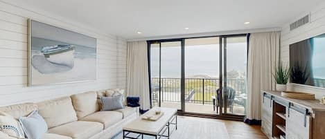 A modern living room with a sofa, coffee table, and a large TV. There is a balcony with a view of the sea.