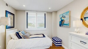 Ein Schlafzimmer mit Bett, einem Fenster mit Jalousien, einem Nachttisch mit Lampe, einem Schminktisch und Kunstwerken an den Wänden.