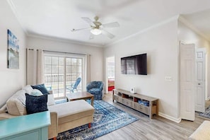 Ein Wohnzimmer mit Sofa, Sessel, Fernseher, Deckenventilator und einem großen Fenster.