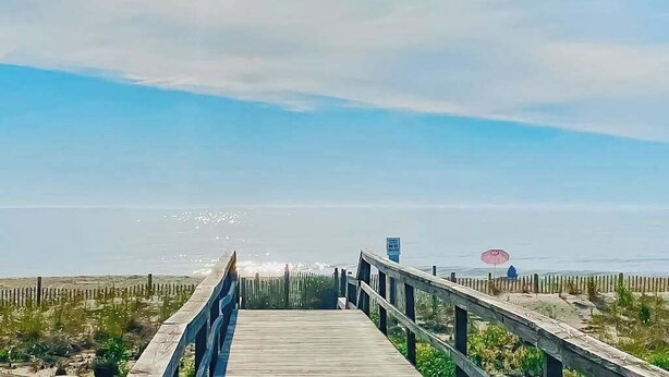 Ein hölzerner Steg führt zum Strand mit freiem Blick auf den Ozean und einem Wegweiser.