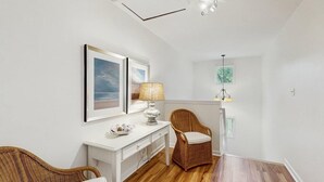 Ein Zimmer mit weißem Schreibtisch, Korbstühlen, einer Lampe und gerahmten Kunstwerken an der Wand.