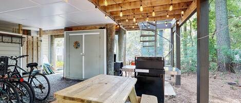 A covered outdoor area with a wooden table and benches, bicycles, and a barbecue grill.
