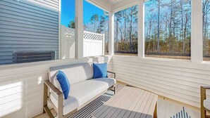 Eine überdachte Veranda mit einem weißen Sofa, blauen Kissen, großen Fenstern und Blick auf Bäume.