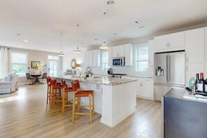 A modern kitchen with white cabinets, a central island with stools, a refrigerator, and a microwave. - Town of Bethany Beach home near beach with firepit, bikes, fireplace, beach gear (Bethany Beach)
