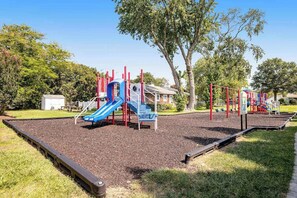 A playground with slides and climbing structures, surrounded by trees and a building in the background. - Bethany West Retreat W/ Shared Pool & Ideal Location Close to Trolley stop (Bethany Beach)