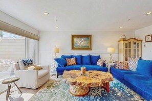 Un salon avec un canapé bleu, un fauteuil blanc, une table basse ronde et un tableau représentant un voilier accroché au mur.
