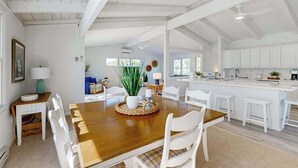 A spacious dining area with a wooden table and white chairs, a kitchen with white cabinets, and a high ceiling with exposed beams. - Ocean Village Beach block, pet friendly house w/patio, balcony, gas grill & WIFI (Bethany Beach)