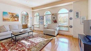 A spacious living room with a sofa, armchair, coffee table, and a TV. There are arched windows with blinds, a ceiling fan, and a large painting of a beach scene. - Town of Ocean View pet friendly house with a private pool! (Bethany Beach)
