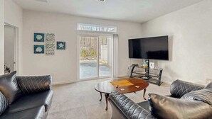 A living room with a flat-screen TV, leather sofas, a coffee table, and a sliding glass door leading to an outdoor area.
