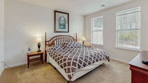 A bedroom with a bed, two bedside tables with lamps, a framed picture on the wall, and two windows with blinds.