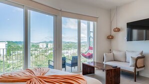 Een moderne hotelkamer met een groot bed, een bank, een hangstoel en een balkon met uitzicht op gebouwen en groen.