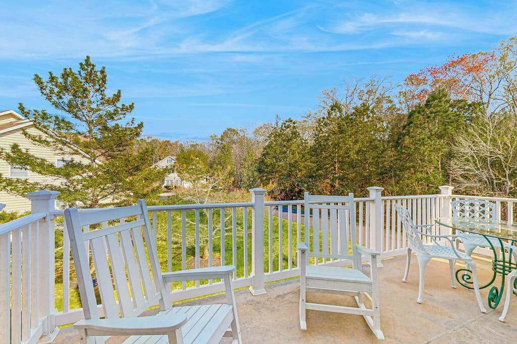 A patio with white wooden chairs and a glass table, surrounded by a white fence and trees.