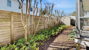 A garden pathway with a wooden fence, a row of young trees, and a building in the background. - Town of Rehoboth Beach - 38242 Robinson Dr. Unit #1 (Rehoboth Beach)