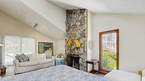 Ein Schlafzimmer mit einem großen Bett, einem Kamin aus Stein, einer Couch und Blick auf Grün durch ein Fenster.