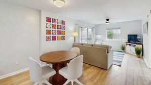 A modern living room with a wooden dining table, white chairs, a beige sofa, and a television on a cabinet. - Henlopen Station condo w/shared pool, patio, grill, close to beach & restaurants (Rehoboth Beach)