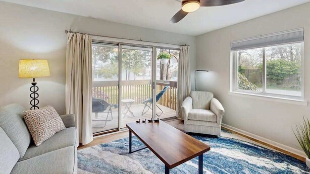 A living room with a sofa, armchair, coffee table, ceiling fan, and large windows. - Henlopen Station condo w/shared pool, patio, grill, close to beach & restaurants (Rehoboth Beach)