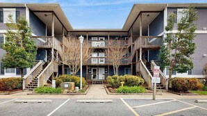 A multi-story apartment building with a central entrance, balconies, and a parking lot. - Henlopen Station condo w/shared pool, patio, grill, close to beach & restaurants (Rehoboth Beach)