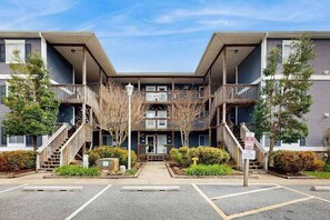 A multi-story apartment building with a central entrance, balconies, and a parking lot. - Henlopen Station condo w/shared pool, patio, grill, close to beach & restaurants (Rehoboth Beach)