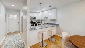 A modern kitchen with white cabinets, a granite countertop, and stainless steel appliances. There are two bar stools and a wooden dining table. - Henlopen Station condo w/shared pool, patio, grill, close to beach & restaurants (Rehoboth Beach)