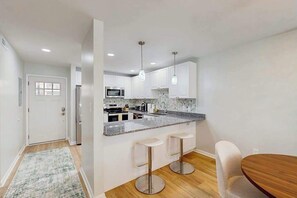 A modern kitchen with white cabinets, a granite countertop, and stainless steel appliances. There are two bar stools and a wooden dining table. - Henlopen Station condo w/shared pool, patio, grill, close to beach & restaurants (Rehoboth Beach)