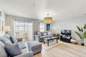 A modern living room with a sofa set, a coffee table, a TV, and a large window with curtains. - The Palms - 36525 Palm Dr, Unit #5201 (Rehoboth Beach)