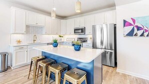Una cocina moderna con armarios blancos, electrodomésticos de acero inoxidable, una isla central con taburetes, iluminación colgante y un elemento decorativo en la pared.
