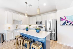 A modern kitchen with white cabinets, stainless steel appliances, a central island with stools, pendant lighting, and a decorative wall piece. - Friendship Creek - 35058 Dundee St | Charming townhouse with pool & patio (Frankford)