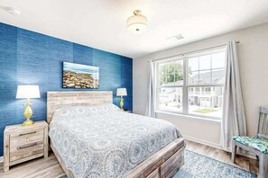 A bedroom with a bed, two bedside tables with lamps, a window with a view, and a blue accent wall. - Friendship Creek - 35058 Dundee St | Charming townhouse with pool & patio (Frankford)