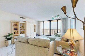 A modern living room with a sofa, a coffee table, and a lamp. There is a bookshelf and a potted plant. - Oceanfront complex with beach access & charm - pools, tennis, fitness centers (Bethany Beach)