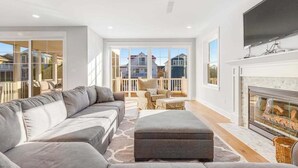 A modern living room with a large grey sectional sofa, a fireplace, and large windows offering a view of neighboring houses. - Town of Bethany Beach home with a fireplace & outdoor living - near beach (Bethany Beach)