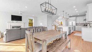 A spacious living area with a wooden dining table, a gray sofa, and a kitchen in the background. - Town of Bethany Beach home with a fireplace & outdoor living - near beach (Bethany Beach)