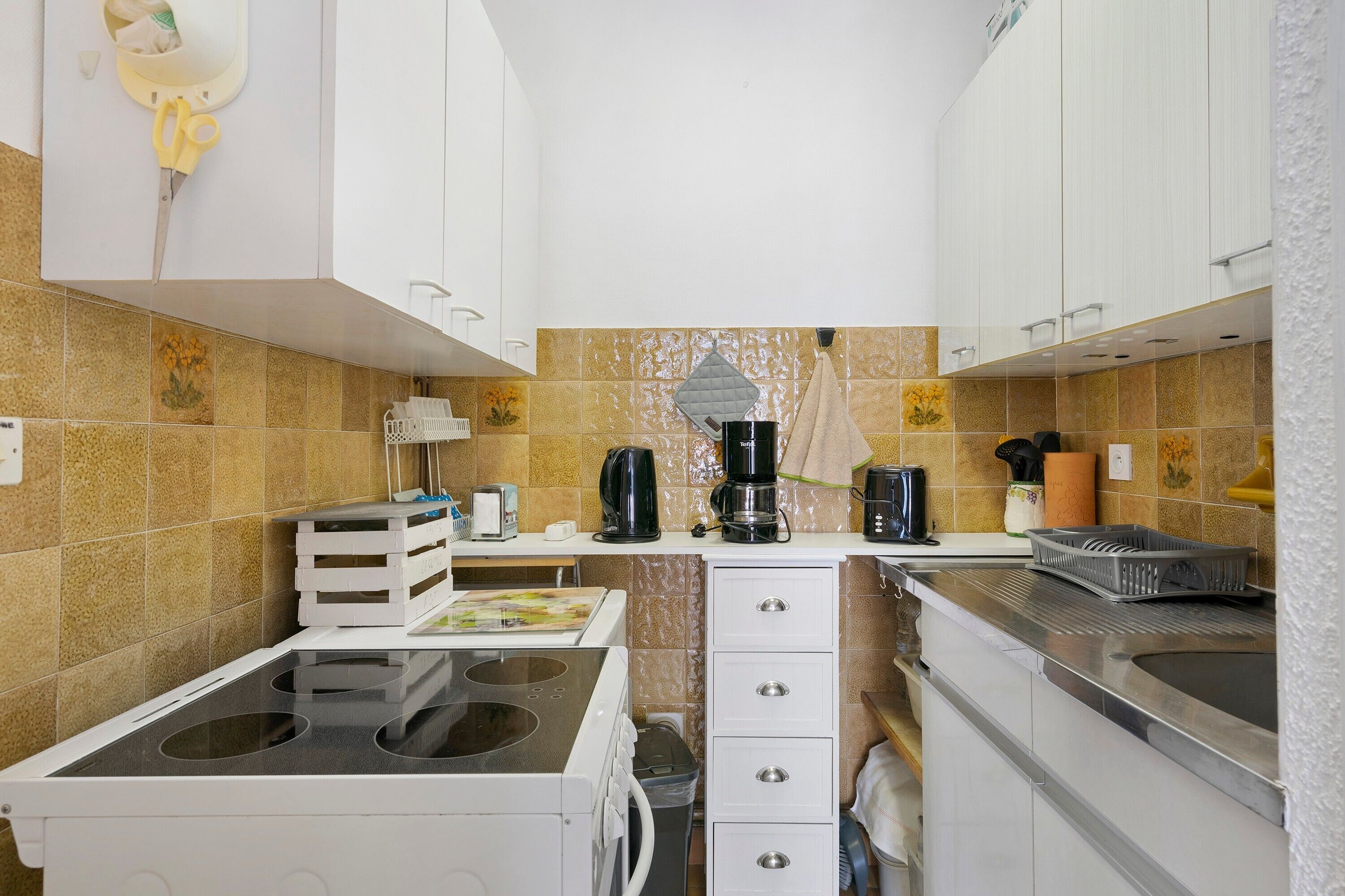 A compact kitchen with a gas stove, white cabinets, a coffee maker, and a trash bin.