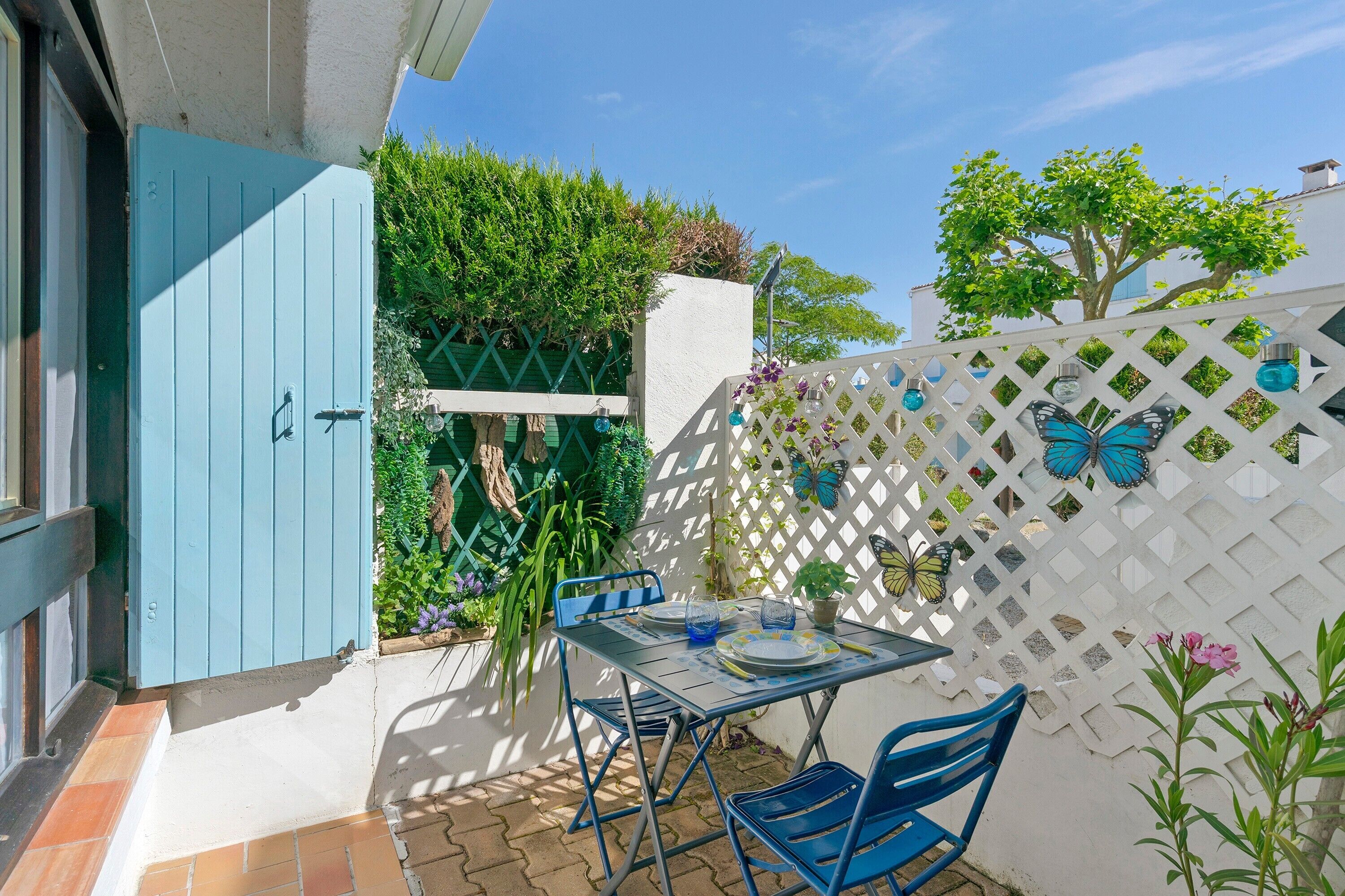 En terrasse med et bord og stole, en blå dør og et hvidt espalier med sommerfugludsmykning.
