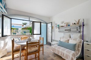 A room with a bed, a table with chairs, a window with a view of buildings, and a shelf with decorative items. - Studio on La Corniche with Shared Pool, Private Terrace, and Shared Garden (Saint-Palais-sur-Mer)