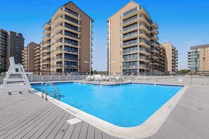 A swimming pool with a wooden deck, surrounded by high-rise residential buildings. - Sea Colony West condo with lovely deck, pools, hot tubs, tennis, & beach shuttle (Bethany Beach)