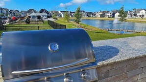 A Blaze gas grill with a digital thermometer, set against a backdrop of a residential area with houses and a body of water.