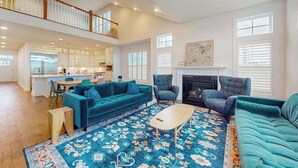 A spacious living area with a sofa, chairs, a coffee table, and a patterned rug. A kitchen is visible in the background, and there is a fireplace with a mantel.
