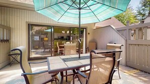 An outdoor patio with a table and chairs, a large umbrella, and a view of the house interior through glass doors. - Experience fun at Sea Colony Bethany Beach - beach, tennis, & pickleball (Bethany Beach)