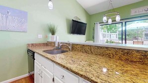 A kitchen with granite countertops, a sink, a potted plant, a flat-screen TV, and a window with a view of greenery. - Experience fun at Sea Colony Bethany Beach - beach, tennis, & pickleball (Bethany Beach)
