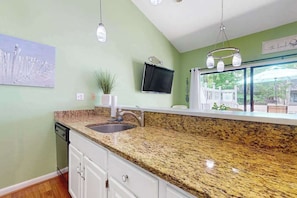 A kitchen with granite countertops, a sink, a potted plant, a flat-screen TV, and a window with a view of greenery. - Experience fun at Sea Colony Bethany Beach - beach, tennis, & pickleball (Bethany Beach)